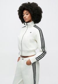 Λευκό τζάκετ με φερμουάρ και ψηλό γιακά, με μαύρες ρίγες τριών γραμμών. Φοριέται με αντίστοιχο λευκό παντελόνι, με άνετη εφαρμογή.