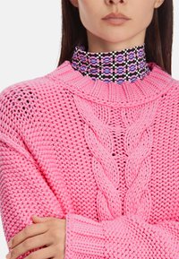 Leuchtend rosa gestrickter Pullover mit Kabelmuster, ausgestattet mit einem hohen Kragen und einem gemusterten Stehkragen in dunklen Tönen mit geometrischen Formen.