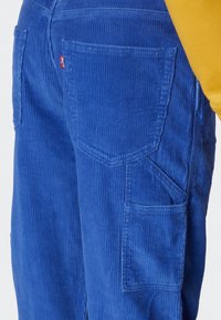 Pantalon en velours côtelé bleu présentant une texture côtelée verticale, des poches arrière avec un détail de couture, et une étiquette de marque visible sur le bord de la poche.