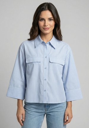 Vrouw met schouderlang donker haar, gekleed in een lichtblauw overhemd met verticale strepen en een lichtblauwe spijkerbroek, staand tegen een effen achtergrond.