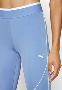 Jasnoniebieskie legginsy wykonane z elastycznego materiału, z białymi szwami i białym logo Pumy na dolnej stronie, o gładkiej fakturze.