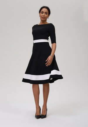 Black midi dress with a fitted top, white waistband, and a flared skirt featuring a white hemline. Worn with black pointed heels.