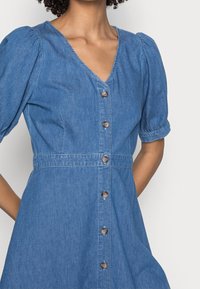 Denimmekko, jossa on V-pääntie, lyhyet puffihihat ja rivi pyöreitä nappeja edessä; vaaleansininen väri ja pehmeä rakenne.