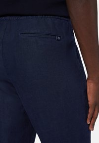 Marinblå linbyxor med en platt bakficka och en liten varumärkesetikett. Tyget har ett texturerat, något skrynkligt utseende.