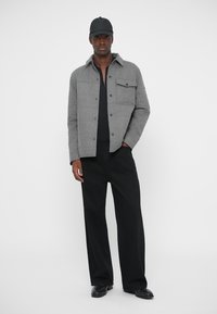 Άνδρας που φοράει γκρι πουκάμισο με κουμπιά, μαύρο πουκάμισο, φαρδιά μαύρα παντελόνια, μαύρο καπέλο και μαύρα παπούτσια, στέκει μπροστά σε λευκό φόντο.