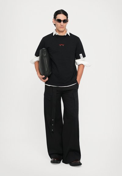 Schwarzes T-Shirt mit rotem Logo, über einem weißen Hemd mit Kragen getragen. Lockere schwarze Cargo-Hosen mit mehreren Taschen, kombiniert mit schwarzem Schuhwerk.