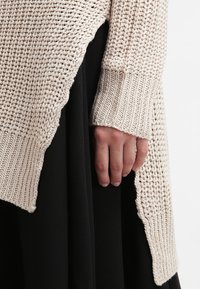 Ręka oparta na boku, ubrana w beżowy gruby sweter z falbankami, na czarnej spódnicy lub spodniach.