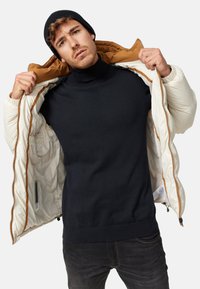 Puffer jacket in off-white with brown lining, unzipped, revealing a dark turtleneck underneath. Model wearing a black beanie, hands holding the jacket.