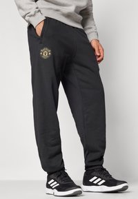 Mustat puuvillaiset collegehousut, joissa on Manchester Unitedin kulta logo, sivutaskut ja joustavat hihansuut. Säädettävä vyötärönauha.