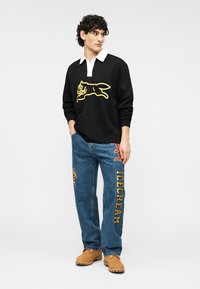 Μαύρο φούτερ με λευκό γιακά, που έχει κίτρινο γραφικό κουνελιού. Ανοιχτό μπλε τζιν παντελόνι με πολύχρωμα μπαλώματα και το κείμενο «ICECREAM». Καφέ υποδήματα.