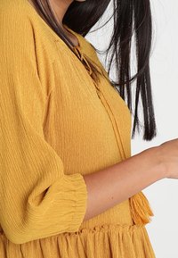 Sininen keltainen mekko teksturoidusta kankaasta, kolmanneksen pituiset hihat ja kerroksellinen hameosa. Koristeena solmuke kaula-aukossa.