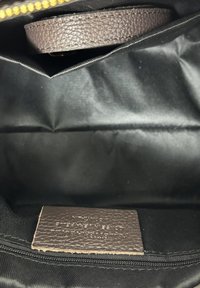 Interno di una borsa di tessuto nero con un'etichetta in pelle marrone testurizzata cucita sopra una tasca con cerniera nera e una cinghia di pelle parzialmente visibile.