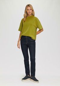 Kurzärmliges, hochgeschlossenes grünes Oberteil aus glattem Stoff, kombiniert mit enganliegenden dunklen Jeans und schwarzen Sneakers mit strukturierten Sohlen.