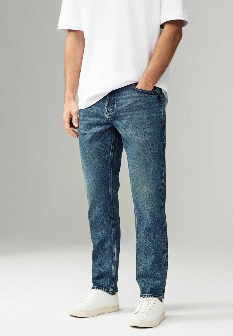 Jeans de denim albastru închis cu o ușoară decolorare, croi drept, cinci buzunare și cusături contrastante, asortați cu adidași albi.