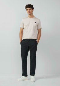 Lichtbeige t-shirt met een ronde hals en klein logo op de borst, gecombineerd met donkere broeken en witte sneakers. Eenvoudig ontwerp en pasvorm.