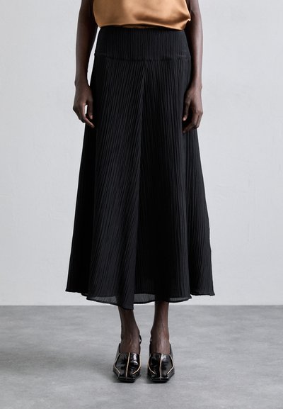 Gonna maxi nera plissettata in tessuto testurizzato, con vita elastica. Indossata con scarpe nere con accenti metallici.