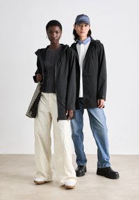 Două modele stau împreună, purtând jachete negre cu glugă. Persoana din stânga are un pulover texturat închis la culoare și pantaloni largi crem, ținând o geantă din plasă. Cealaltă persoană poartă o cămașă albă cu guler bleu deschis, blugi și pantofi chunky negri.