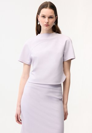 Ung kvinde iført lyse lavendel kortærmet top og matchende nederdel, stående mod en ensfarvet hvid baggrund, vendt fremad.