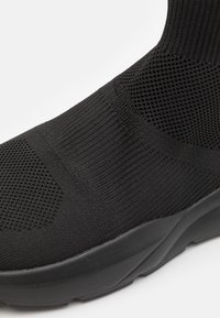 Black knitted sneaker with a textured upper, featuring a smooth sole and elastic ankle design for snug fit and breathability.