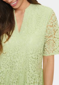 Fényzöld csipkeruha V-kivágással és rövid csipkeujjakkal, virágmintákkal díszítve és bővülő szoknyával.