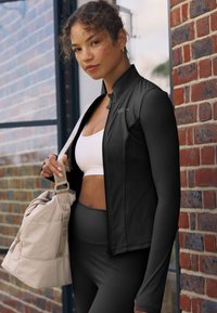 Zwarte aansluitende sportjas met lange mouwen, gedragen over een witte cropped top en gecombineerd met donkere leggings. Beige sporttas in één hand gehouden.