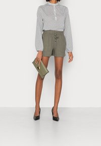 Mustavalkoinen pilkullinen paita, pitkät ilmapallohiha, yhdistetty oliivinvihreisiin nyörillä kiristettäviin shortseihin. Pitää kädessään oliivinvihreää kirjekuorilaukkua, päällä mustat korkokengät.