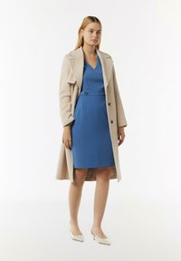 Blaues Kleid mit V-Ausschnitt und tailliertem Schnitt, unter einem beigen Übermantel getragen. Das Model trägt weiße Absätze, und der Stoff wirkt glatt.