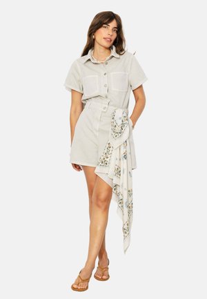 Femme portant une robe beige à manches courtes avec boutons, deux poches poitrine, une ceinture foulard fleurie, un collier de perles et des sandales marron.