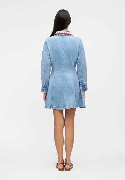 Γυναίκα που στέκεται γυρισμένη πλάτη, φοράει ανοιχτό μπλε φόρεμα από τζιν με μακριά μανίκια και κόκκινο γιακά με κυματιστό τελείωμα, και καφέ σανδάλια.