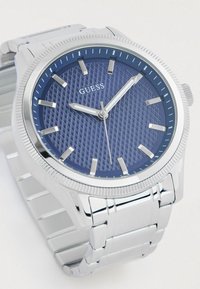 Montre en acier inoxydable avec un cadran texturé bleu, des index heures en argent et un bracelet poli. Présente une lunette striée et un logo.