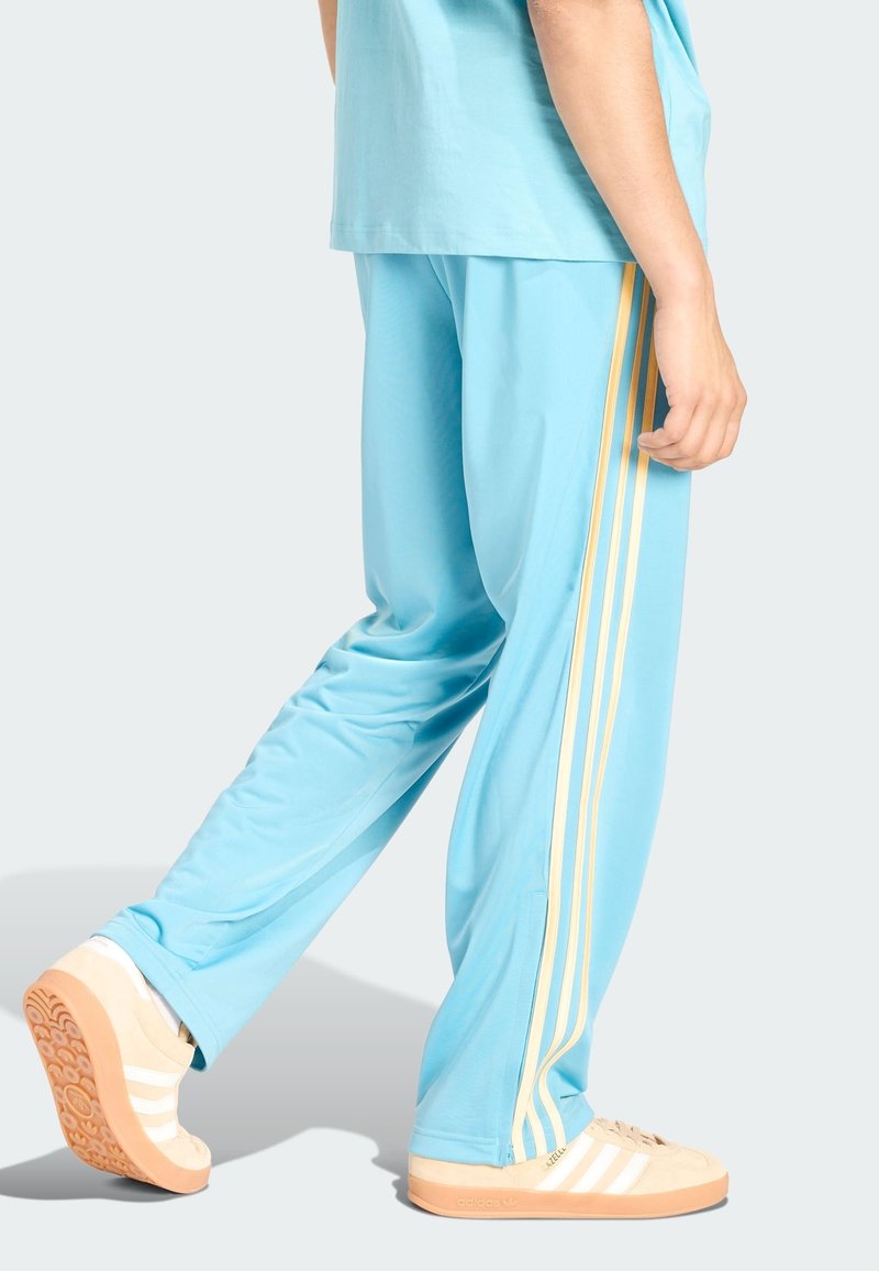 Persona con pantaloni della tuta azzurri chiaro con tre strisce beige sul lato e sneakers Adidas beige con suole di gomma, in piedi di lato.