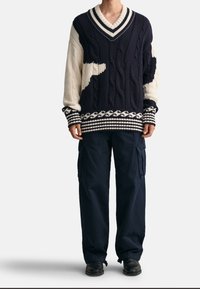 Námořnický a krémový oversize pletený svetr se vzorem copánků, výstřihem do V a proužkovanými manžetami, v kombinaci s námořnickými cargo kalhotami a černými botami.