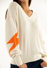 Maglione color crema con scollo a V e maniche lunghe. Presenta un design a forma di fulmine arancione sulla manica sinistra e polsini a coste.