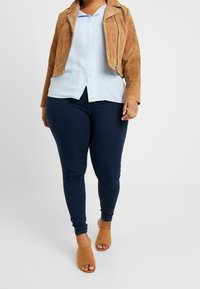 Giacca in suede marrone sopra una camicia azzurro chiaro con bottoni, abbinata a jeans skinny blu scuro e mule beige senza tacco. Design semplice e sartoriale.