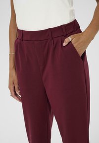Pantalons taille haute bourgogne avec poches latérales, ajustement sur mesure et plis subtils. Le matériau semble lisse avec un léger éclat.