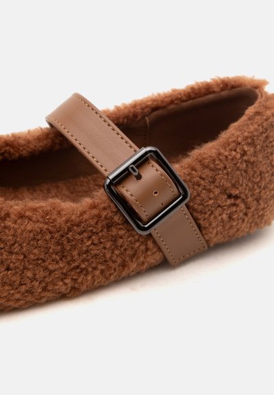 Chaussure plate marron duveteuse avec une large sangle en cuir et une boucle carrée noire sur fond blanc.