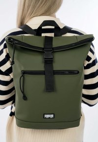 Sac à dos à rabat vert fabriqué en matériau imperméable, doté de bretelles noires réglables, d'une poche zippée à l'avant et d'une étiquette à code-barres au fond.