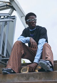 Black t-shirt layered over a light blue long-sleeve shirt, brown checked wide-leg pants, sunglasses, and black sneakers on metal stairs.