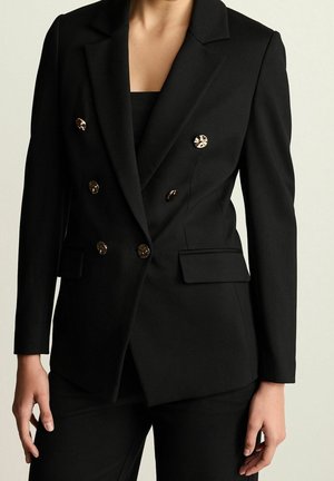 Femme portant un blazer noir croisé à boutons dorés et un pantalon noir assorti, debout devant un fond uni.