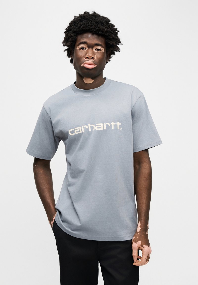 Grijs katoenen t-shirt met korte mouwen en ronde hals, met het witte "carhartt." logo aan de voorkant. Soepele textuur en comfortabele pasvorm.