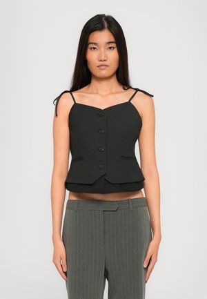Mujer vestida con una blusa sin mangas de botones negra con tirantes atados y pantalones de rayas grises, de pie contra un fondo sencillo.