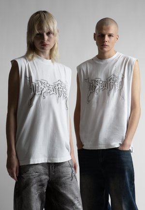 Δύο νέοι ενήλικες που φορούν λευκά αμάνικα μπλουζάκια με κείμενο σε στυλ γκράφιτι, στέκονται δίπλα-δίπλα μπροστά σε απλό φόντο.