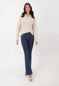 Cremfarbener, gerippter Strickpullover mit einem Viertelreißverschluss, kombiniert mit dunkelblauen Schlagjeans und weißen offenen High Heels auf einem schlichten Hintergrund.