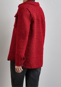 Red textured jacket with a loose fit, featuring a collar, two chest pockets, and black button accents. Material appears woven and chunky.