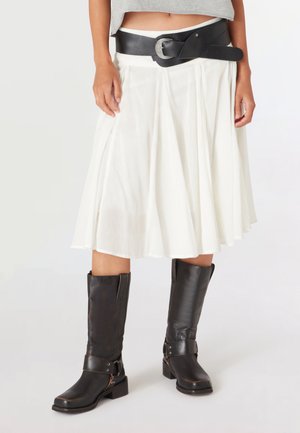 Mujer con falda blanca hasta la rodilla, cinturón ancho negro y botas de piel negras hasta la mitad de la pantorrilla sobre un fondo liso.