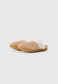 Et par beige slip-on tøfler med hvit, fluffy fôr og gummisåle på en ensfarget lys bakgrunn.