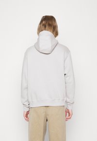 Ljusgrå hoodie med stor luva, ribbad fåll och ärmslut, tillverkad av mjukt material. Vy bakifrån som visar söm detaljer.