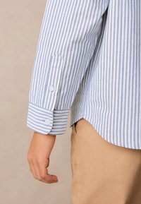 Kék-fehér csíkos hosszú ujjú ing, gombos mandzsettával, könnyű anyagból, világos bézs nadrágba behúzva.