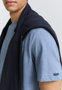 Tricou ribbed de un albastru deschis, cu un pulover tricotat de un bleumarin închis aruncat pe umăr, având un eticheta cu logo pe mânecă.