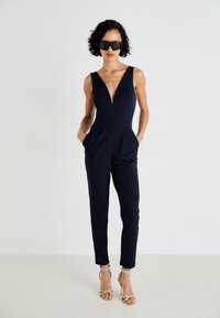 Ολόσωμη φόρμα σε ναυτικό μπλε χρώμα χωρίς μανίκια, με βαθύ V-λαιμό, πλαϊνές τσέπες και κωνικά μπατζάκια, συνδυασμένη με χρυσά πέδιλα με λουράκια. Λεία υφή.